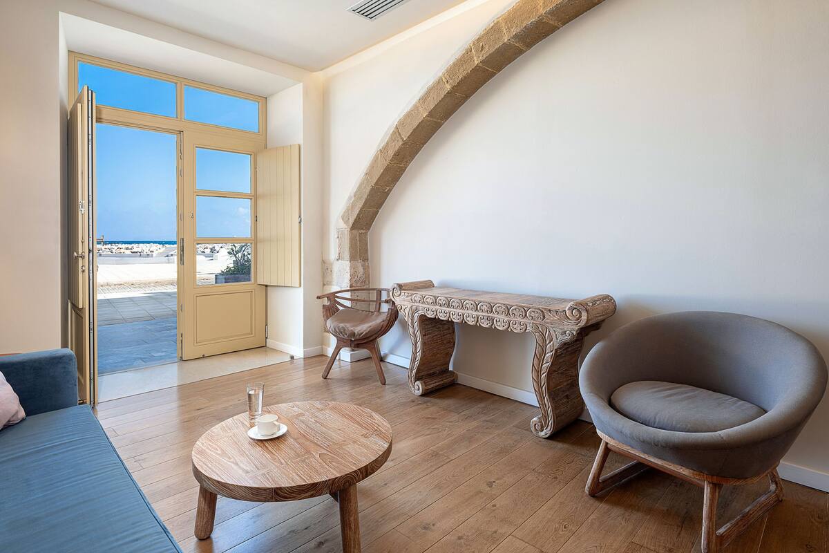 A room with a wooden floor, a round wooden table with a cup and saucer, a grey armchair, a bench with intricate carvings, and a door leading to a balcony with a view of the sea.