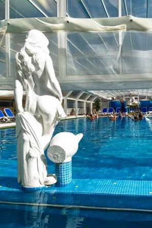 A swimming pool with a white statue, clear blue water, and a covered area with blue chairs.