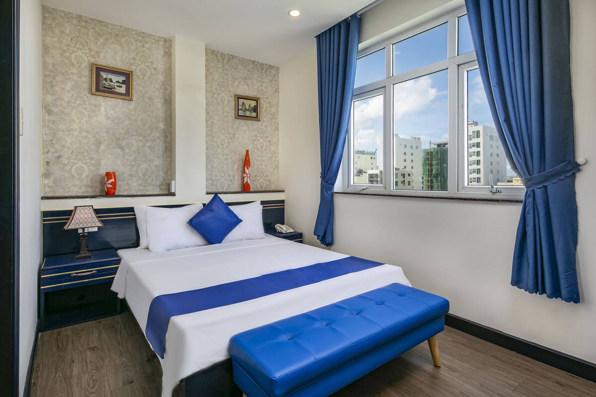 A hotel room with a large bed, a blue bench, a nightstand with a lamp, and a window with blue curtains.