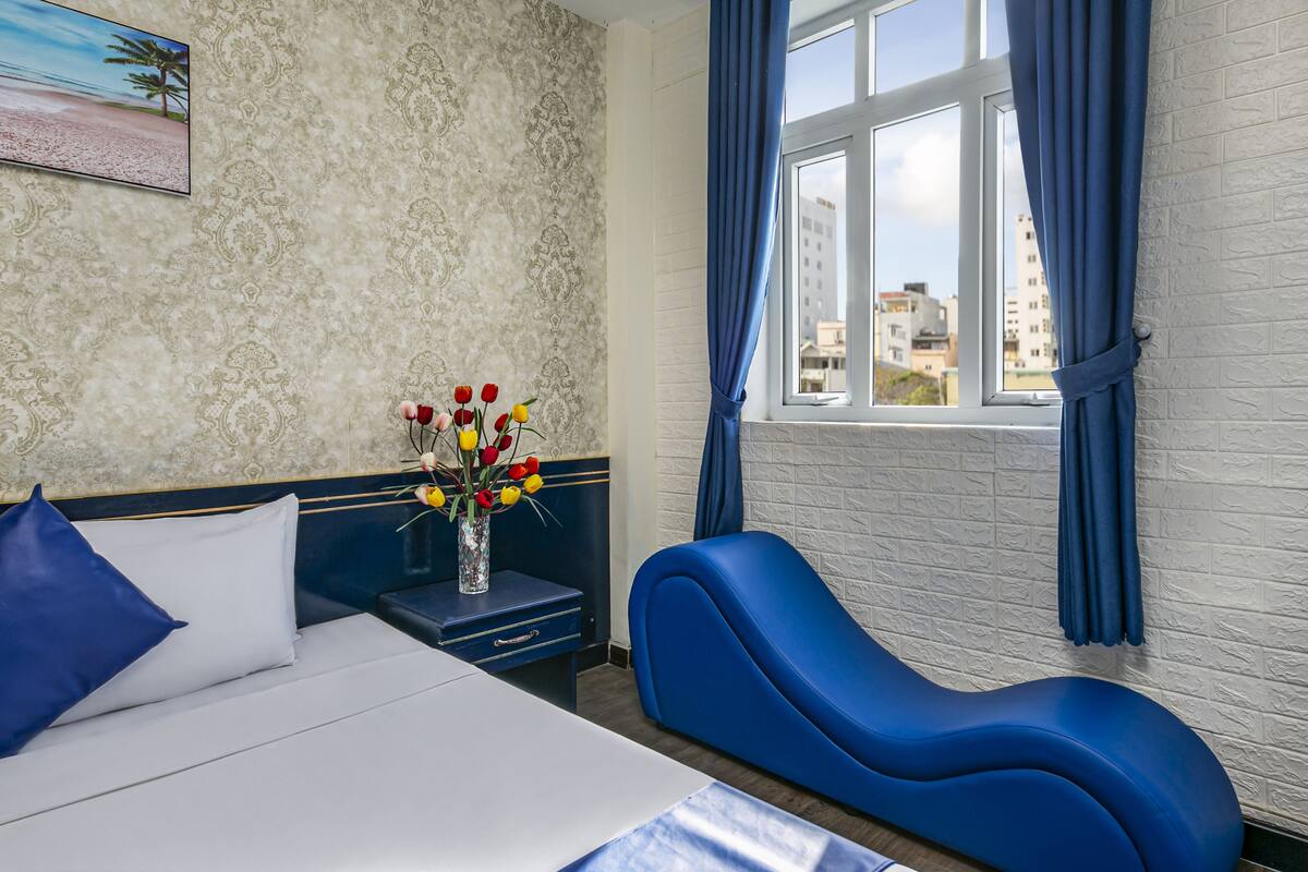 A bedroom with a bed, a nightstand, a vase with flowers, a window with curtains, and a view of buildings outside.