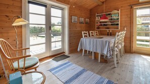Interior de uma cabana de madeira com uma mesa de jantar, cadeiras, um abajur e uma estante de livros.