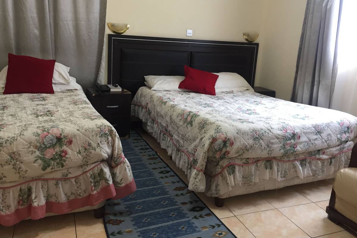 A hotel room with two beds, each with floral bedding and red pillows. There is a nightstand with a phone and a book, and a blue patterned rug on the floor.