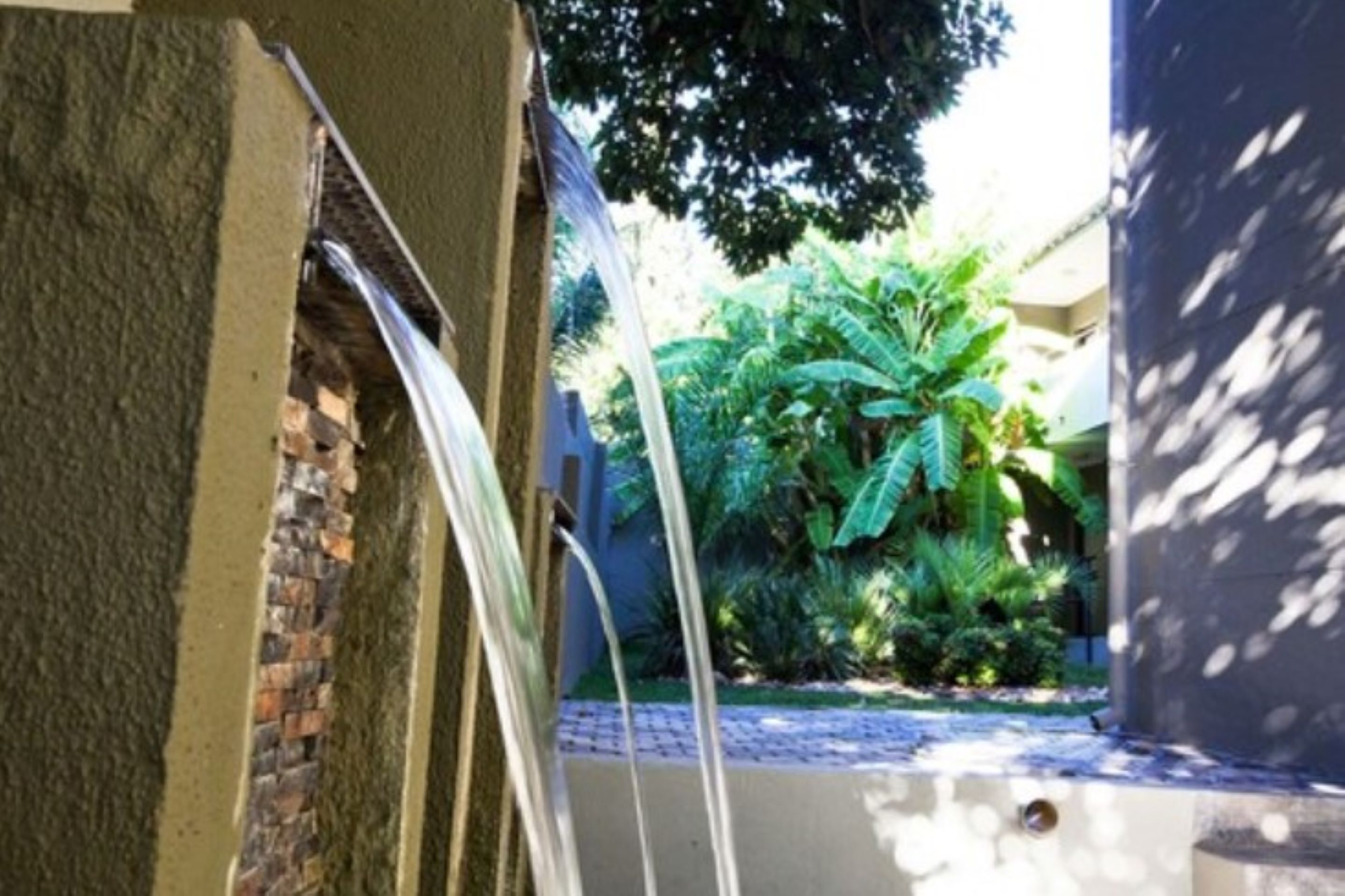 Un estrecho pasillo exterior con un muro de ladrillo en un lado y un muro de hormigón en el otro, que conduce a una exuberante zona ajardinada con diversas plantas y árboles.