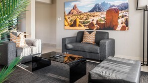 A living room with two grey sofas, a coffee table, and a large desert landscape painting on the wall. - Hillside Executive Accommodation (Windhoek)