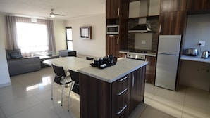 A modern kitchen with a central island, dark wood cabinets, and stainless steel appliances. There is a dining area with a glass table and chairs, and a living room with a sofa and a television. - Hillside Executive Accommodation (Windhoek)