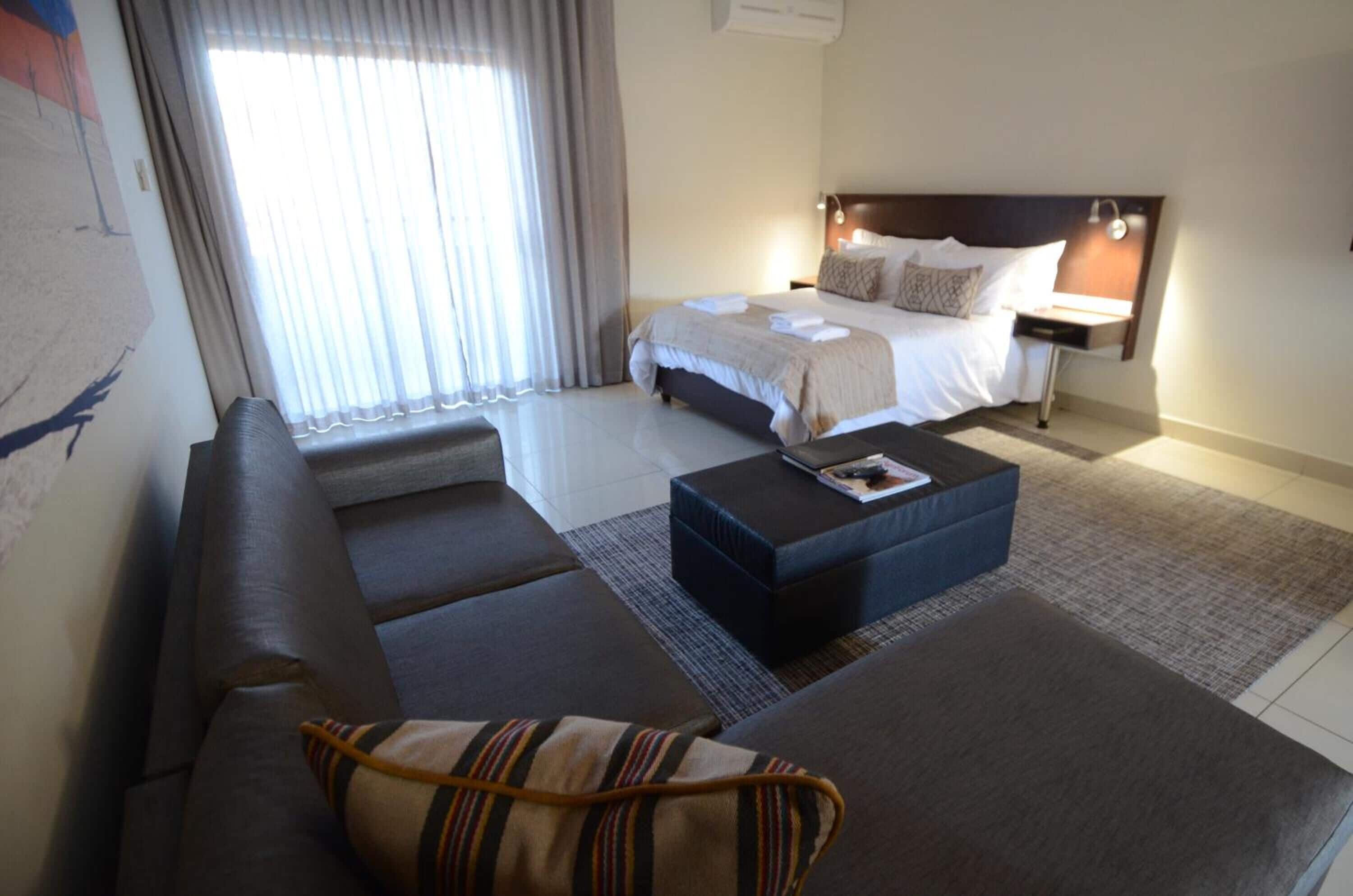 A hotel room with a large bed, a sofa, a coffee table, and a nightstand with a lamp.