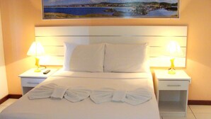 A hotel room with a neatly made bed, two bedside tables with lamps, a painting above the bed, and a view of a coastal landscape. - Pousada Águas do Forte (Cabo Frio)