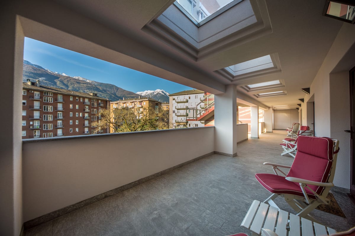 A balcony with a view of mountains and buildings, featuring a row of red lounge chairs and a skylight.