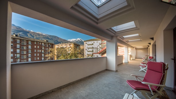 A balcony with a view of mountains and buildings, featuring a row of red lounge chairs and a skylight. - Piazzi House (Sondrio)