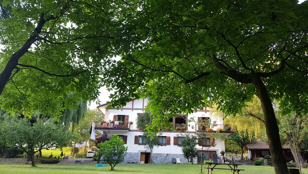 Una casa de dos pisos con balcón, rodeada de un césped cuidado y árboles.