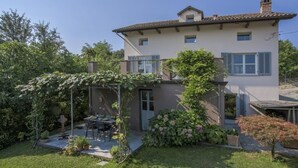 Ein zweigeschossiges Haus mit überdachter Terrasse, einem Garten mit verschiedenen Pflanzen und einer kleinen Sitzgelegenheit im Freien.