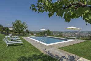 Ein rechteckiger Pool mit glasklar blauem Wasser, umgeben von einer gepflasterten Liegefläche. Rund um den Pool stehen Liegestühle und Sonnenschirme, im Hintergrund ist eine ferne Stadt und Hügellandschaft zu sehen.