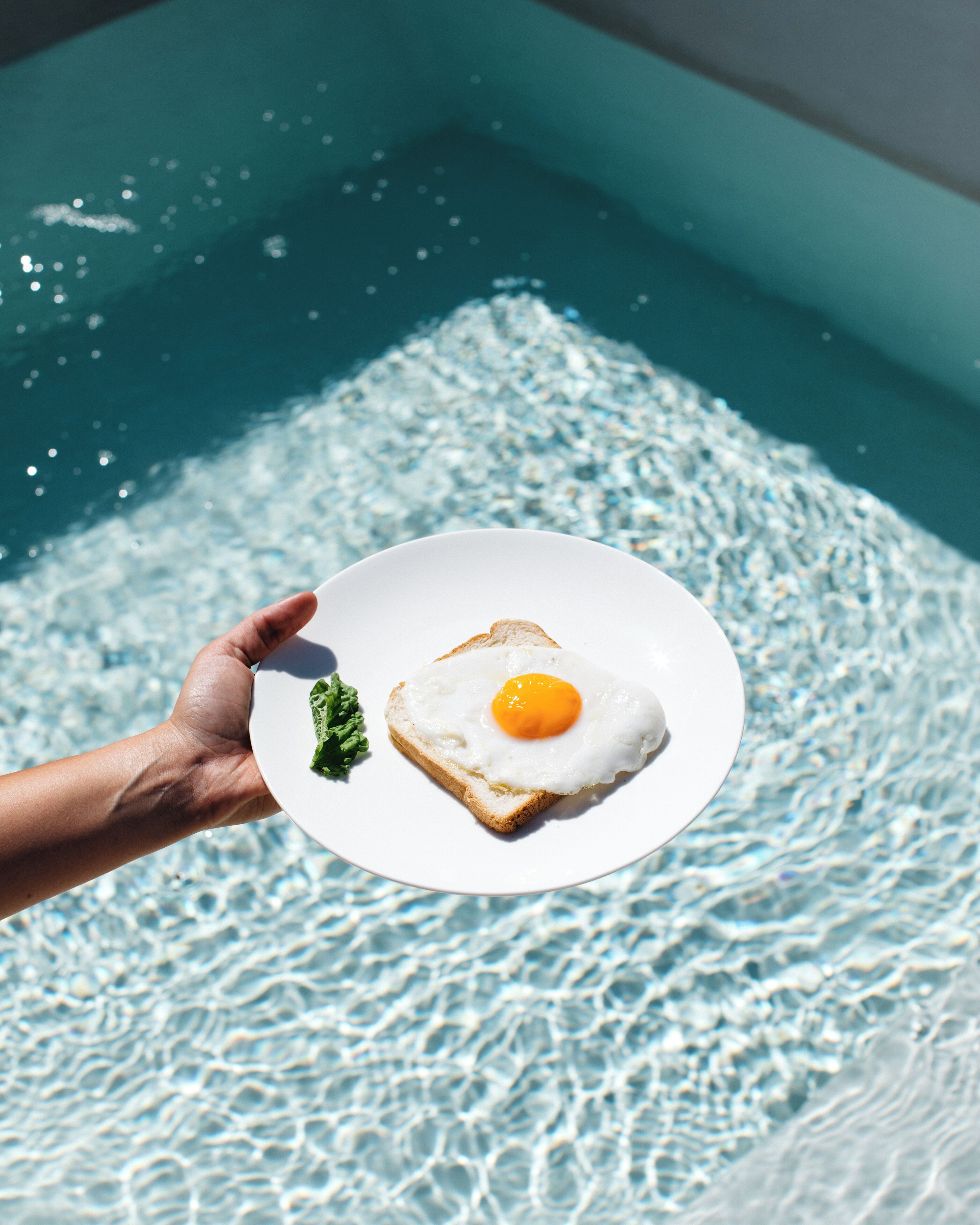En hånd holder en tallerken med spejlæg og en skive ristet brød over en pool.