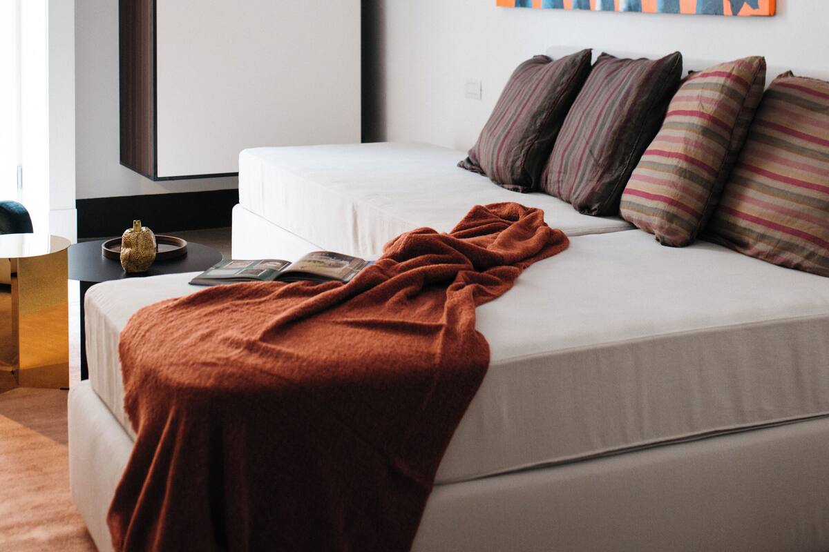 A modern bedroom with a bed, a bedside table, a wall-mounted shelf, and a large abstract painting.