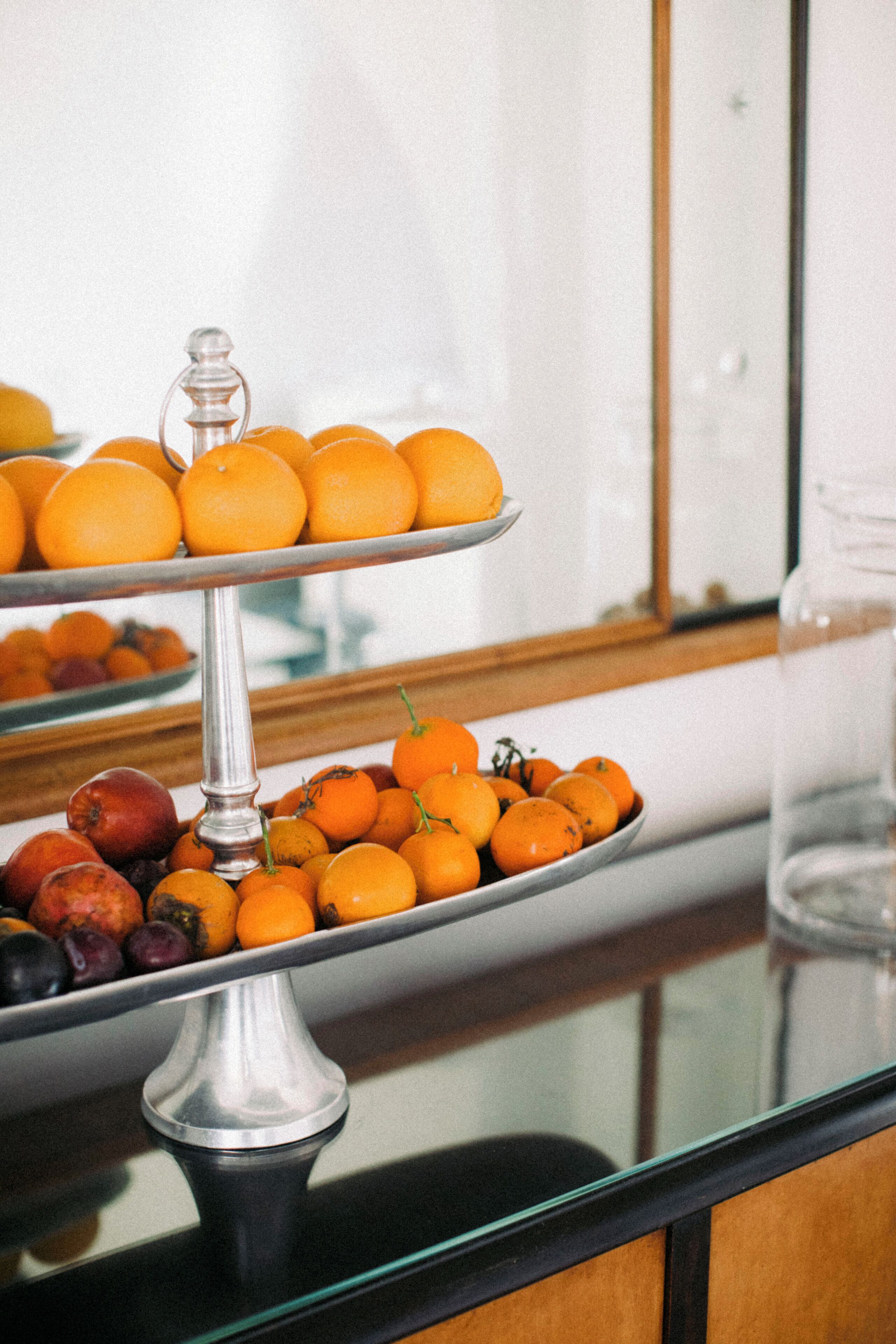A three-tiered fruit stand with oranges and other fruits on a glass table.