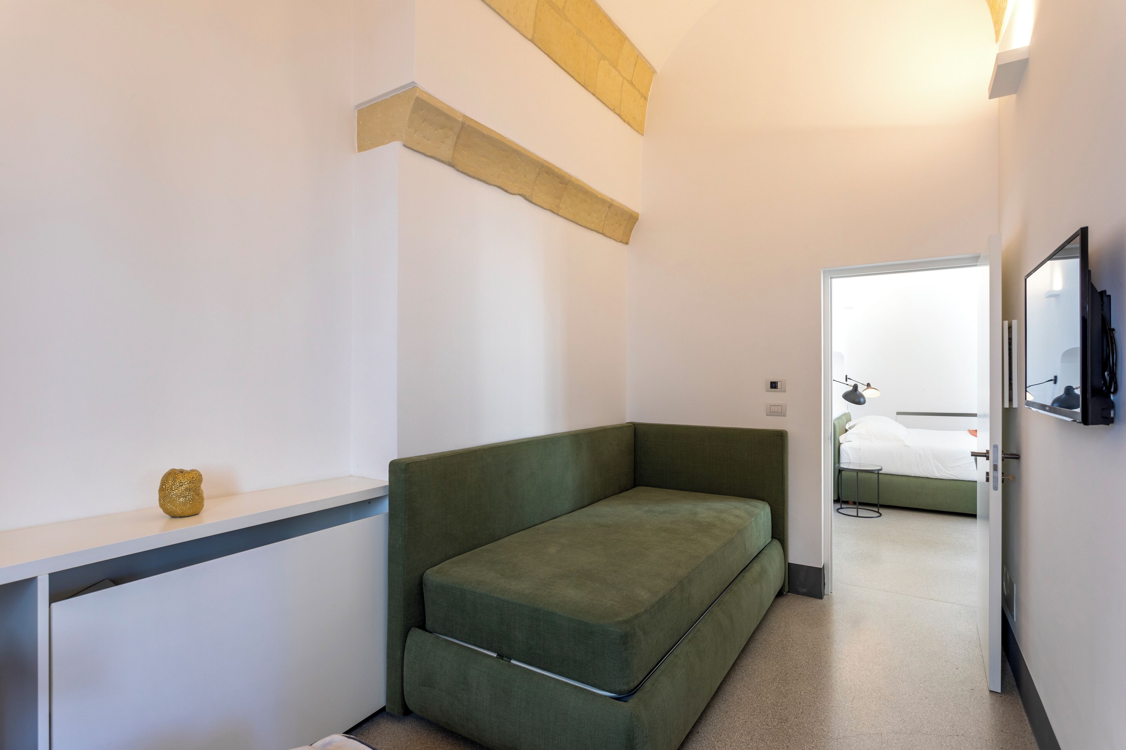 Una habitación moderna y compacta con un sofá verde, una mesita pequeña y una cama al fondo.