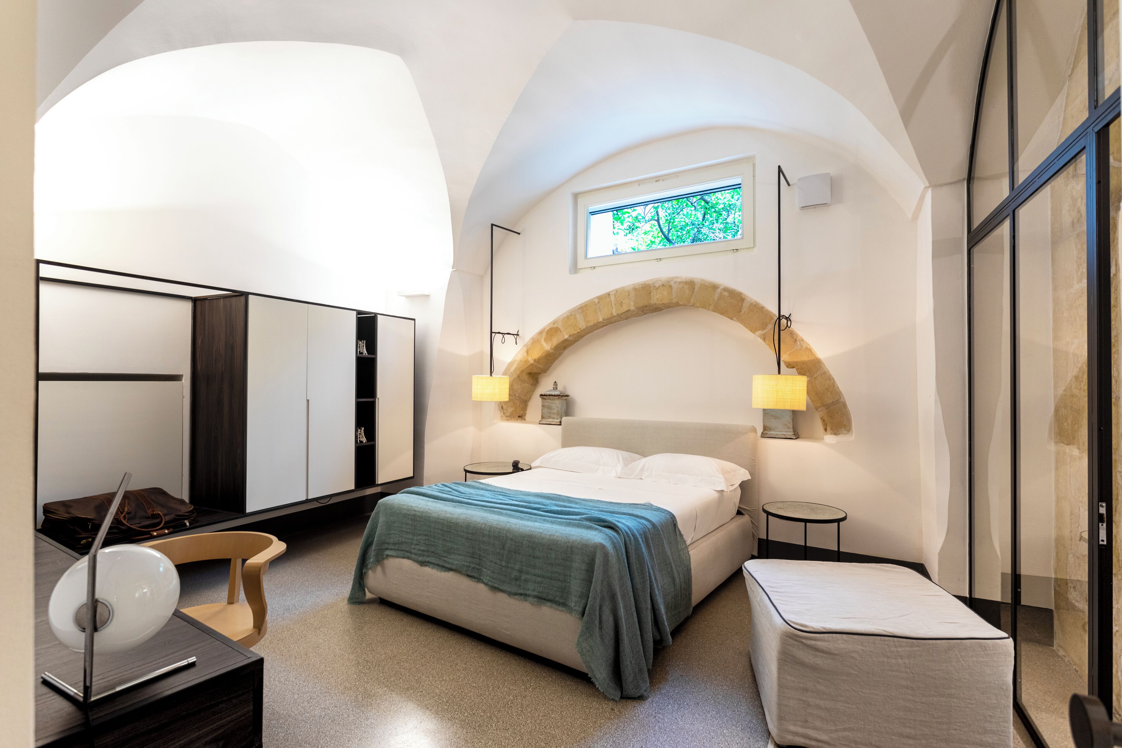 Un dormitorio moderno con un arco de piedra curvado, una cama con manta verde, un escritorio con una computadora portátil y una mesita de noche con lámpara.