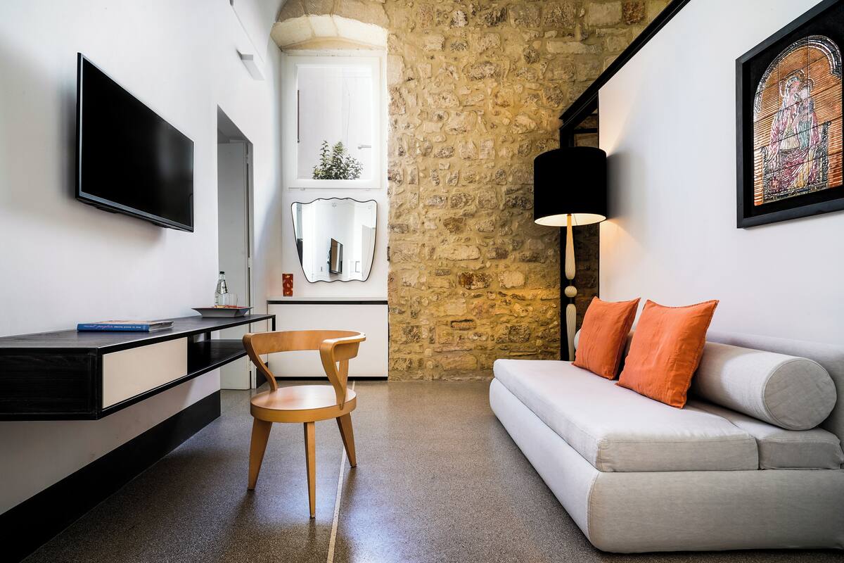 A modern living room with a stone wall, a flat-screen TV, a wooden chair, a sofa with orange pillows, and a framed artwork.