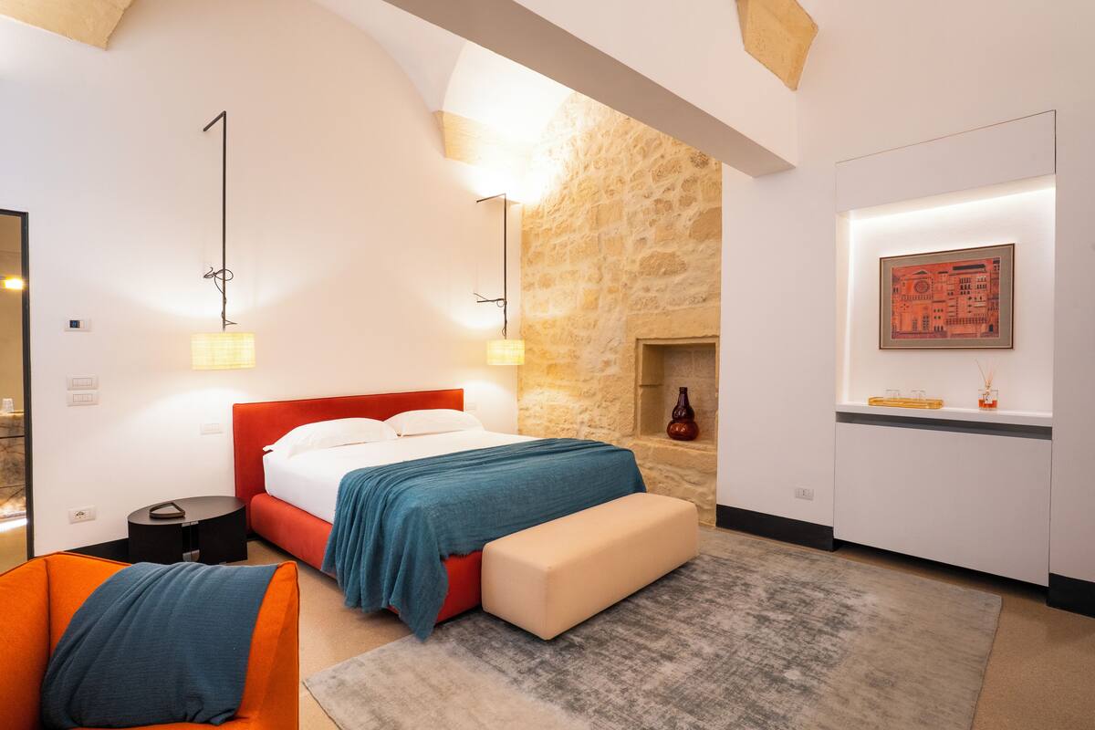 A modern hotel room with a stone accent wall, a large bed with a red headboard, a bench, a small table, and a framed picture on the wall.