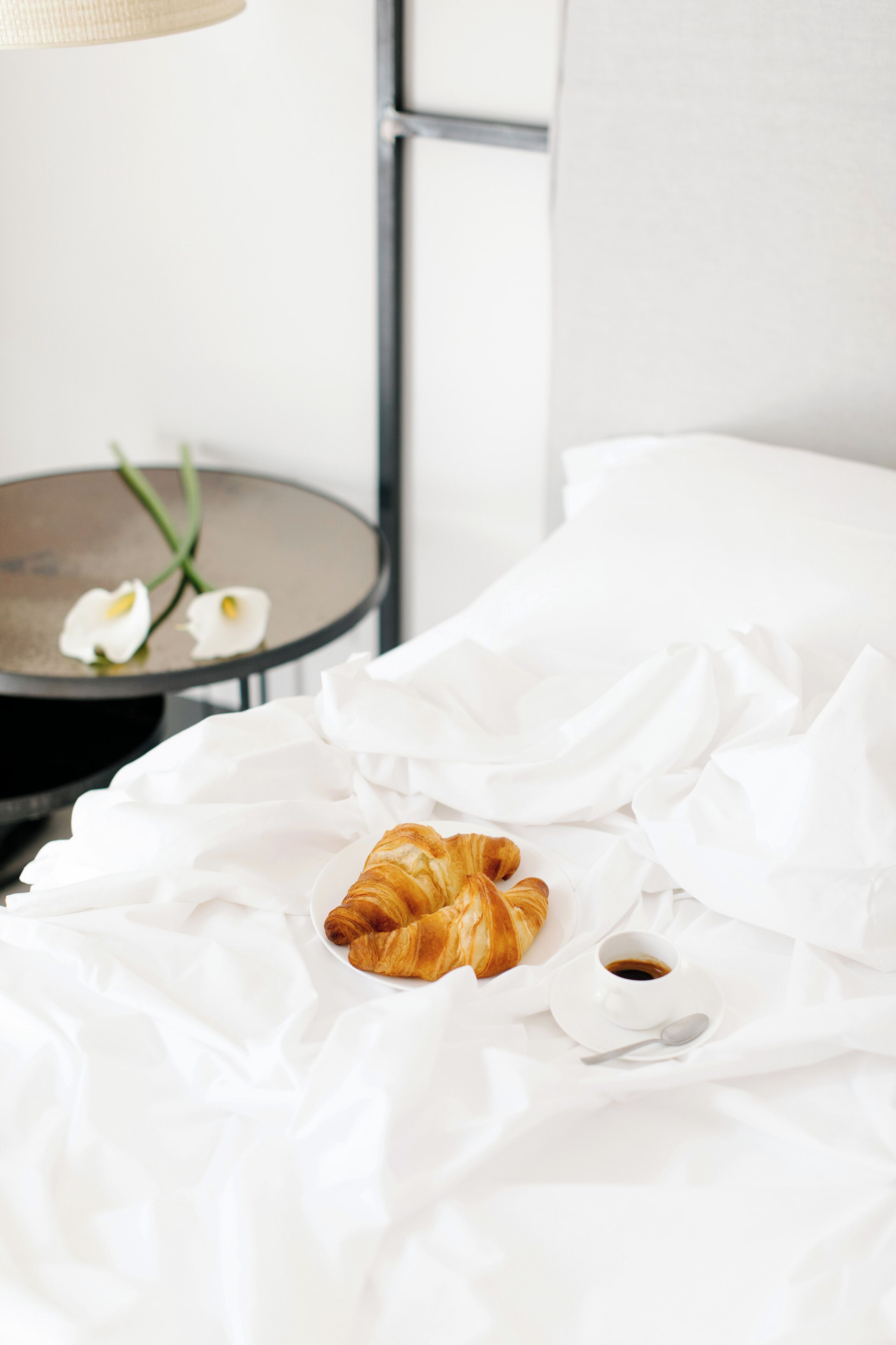 A neatly made bed with white linens, a croissant, and a cup of coffee on a bedside table.