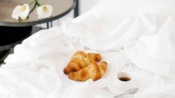 A neatly made bed with white linens, a croissant, and a cup of coffee on a bedside table.