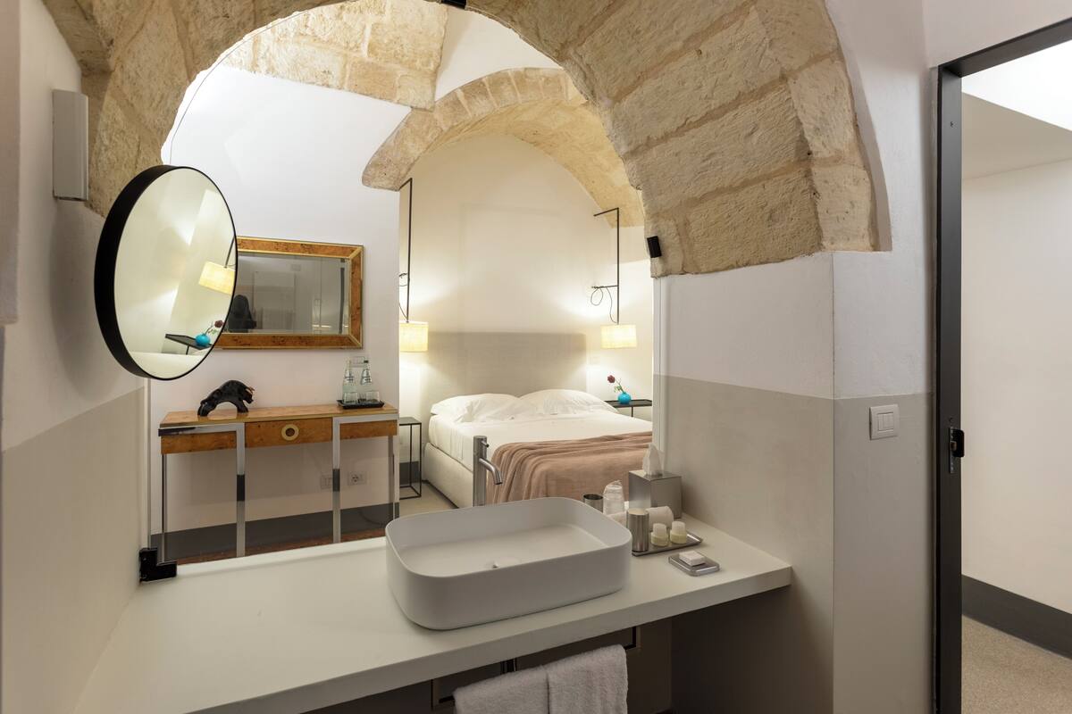 A modern bathroom with a stone arch, a large mirror, a rectangular sink, and a bedside table with a lamp.