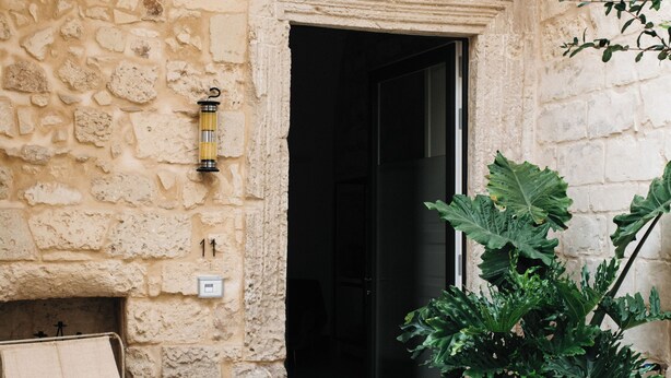 A stone building entrance with a metal chair and a large potted plant. - Palazzo De Noha (Lecce)