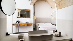 A modern bathroom with a large mirror, a wooden vanity, a bed, and a stone archway. - Palazzo De Noha (Lecce)