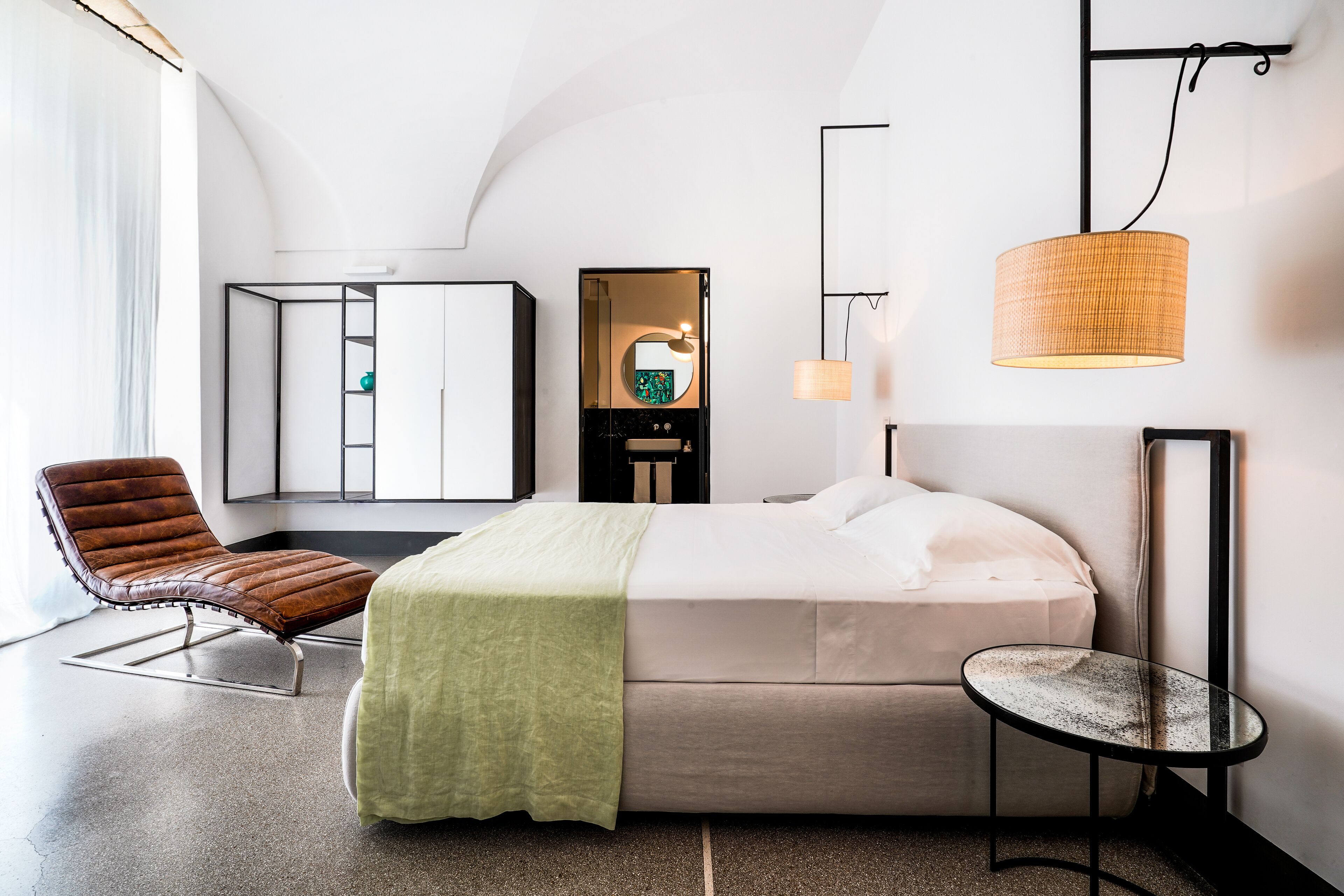 Moderni hotellihuone, jossa on sänky, tuoli, pieni pöytä, peili ja riippuvalaisin.