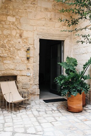 A stone building entrance with a door, a metal chair, and a large potted plant. - Palazzo De Noha (Lecce)