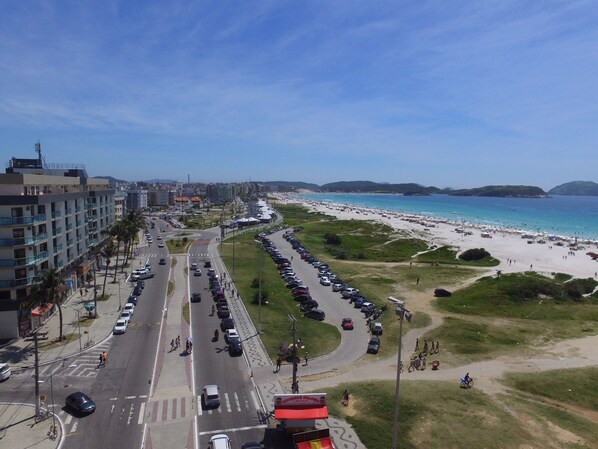 Uma estrada costeira com carros estacionados, uma praia e um hotel com varandas com vista para a praia.