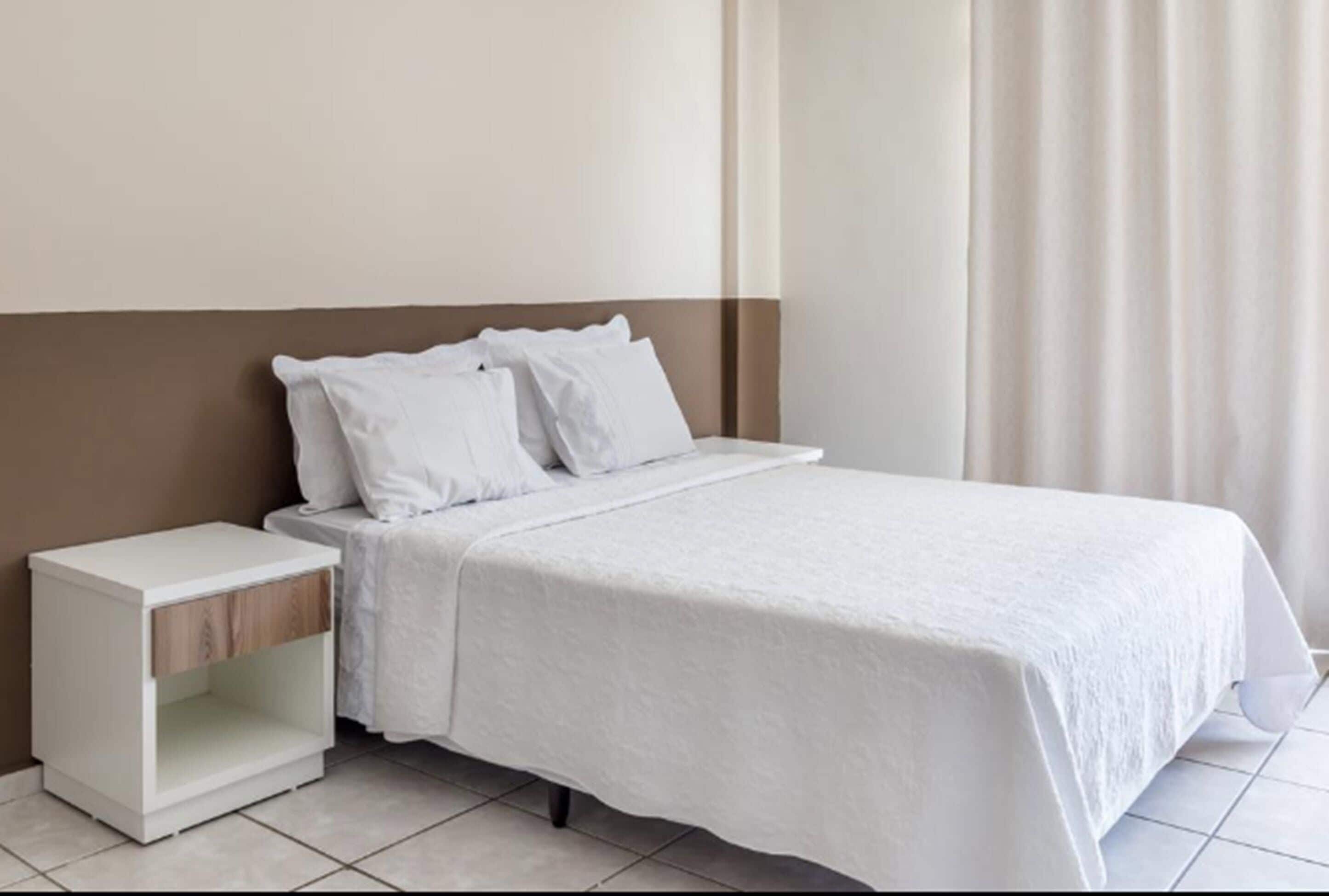 A neatly made bed with white bedding, a nightstand with a drawer, and a plain wall with a curtain.