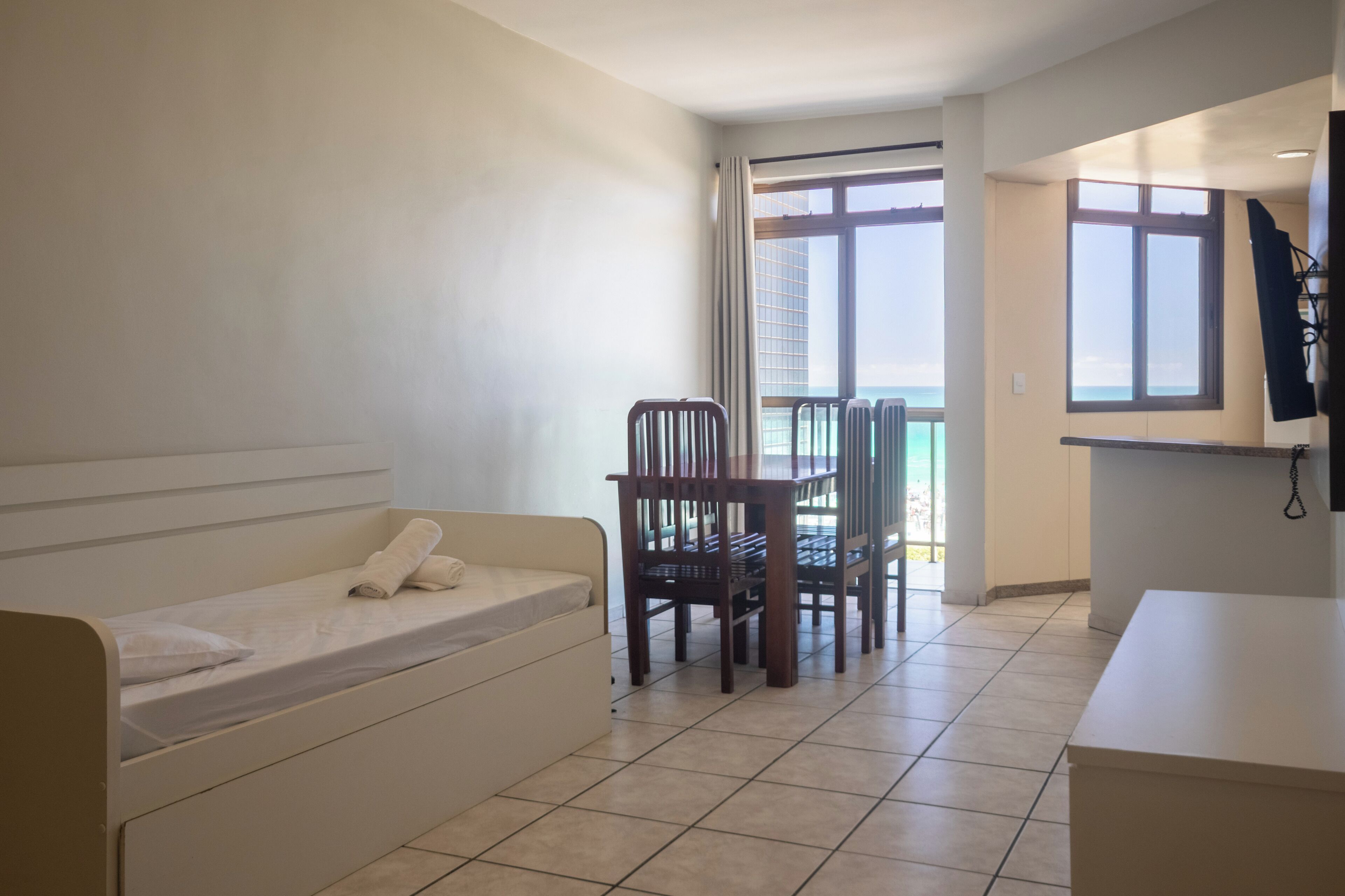 Ein kleiner, sauberer Raum mit einem Bett, einem Holzstuhl, einem kleinen Tisch und Blick auf das Meer durch große Fenster.