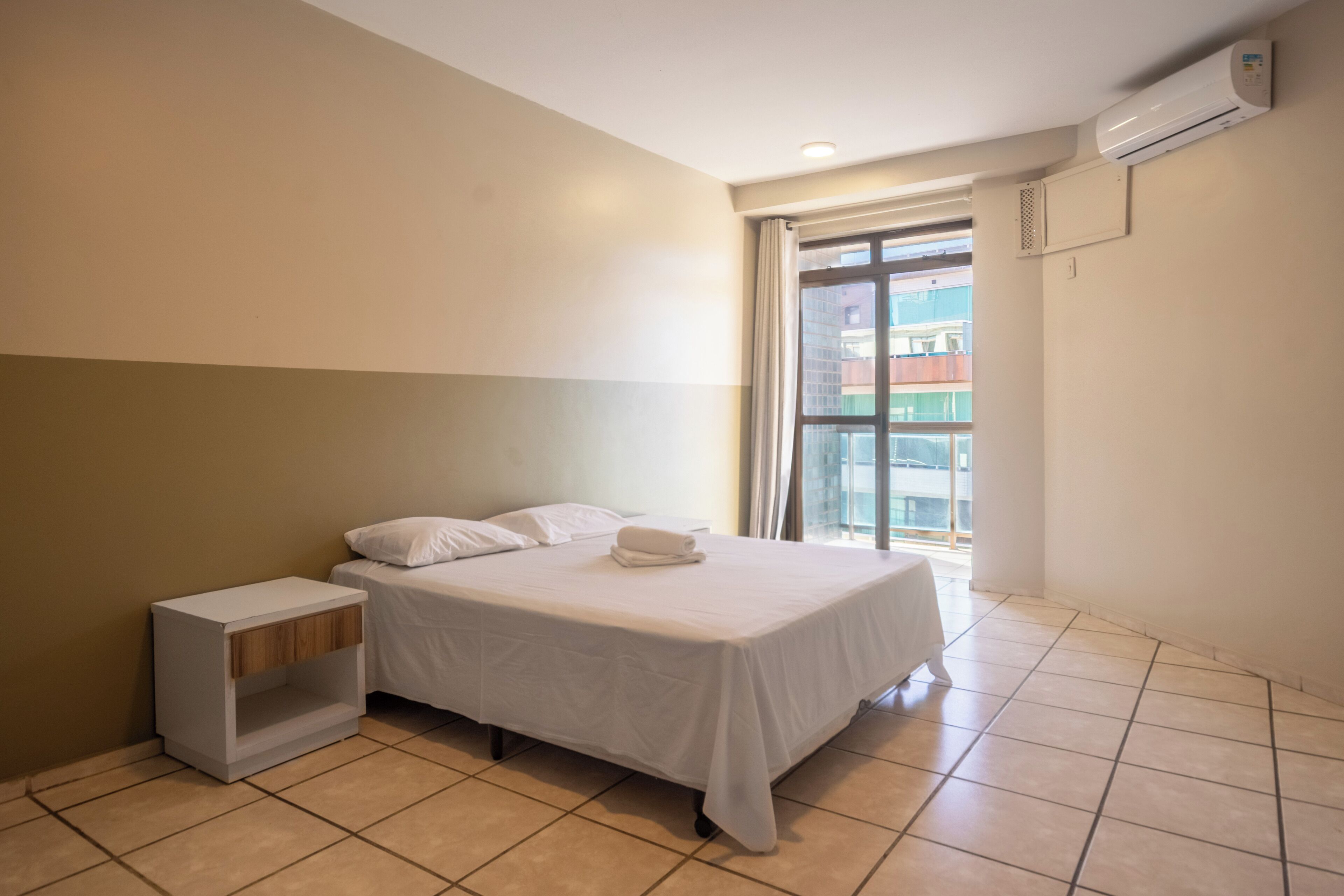 A bedroom with a bed, a nightstand, a window, and an air conditioning unit.