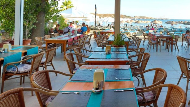 An outdoor dining area with wicker chairs and tables, overlooking a beach and marina. - Gumbet Anil Beach (Bodrum)