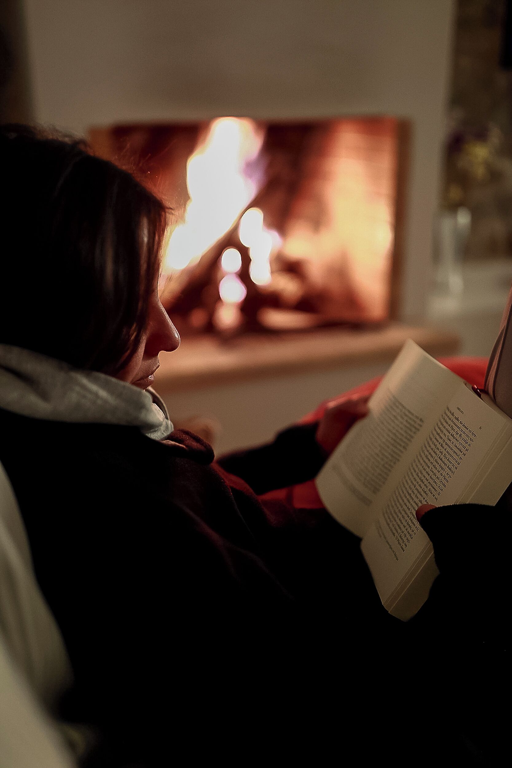 A person reading a book in a dimly lit room with a fireplace in the background.