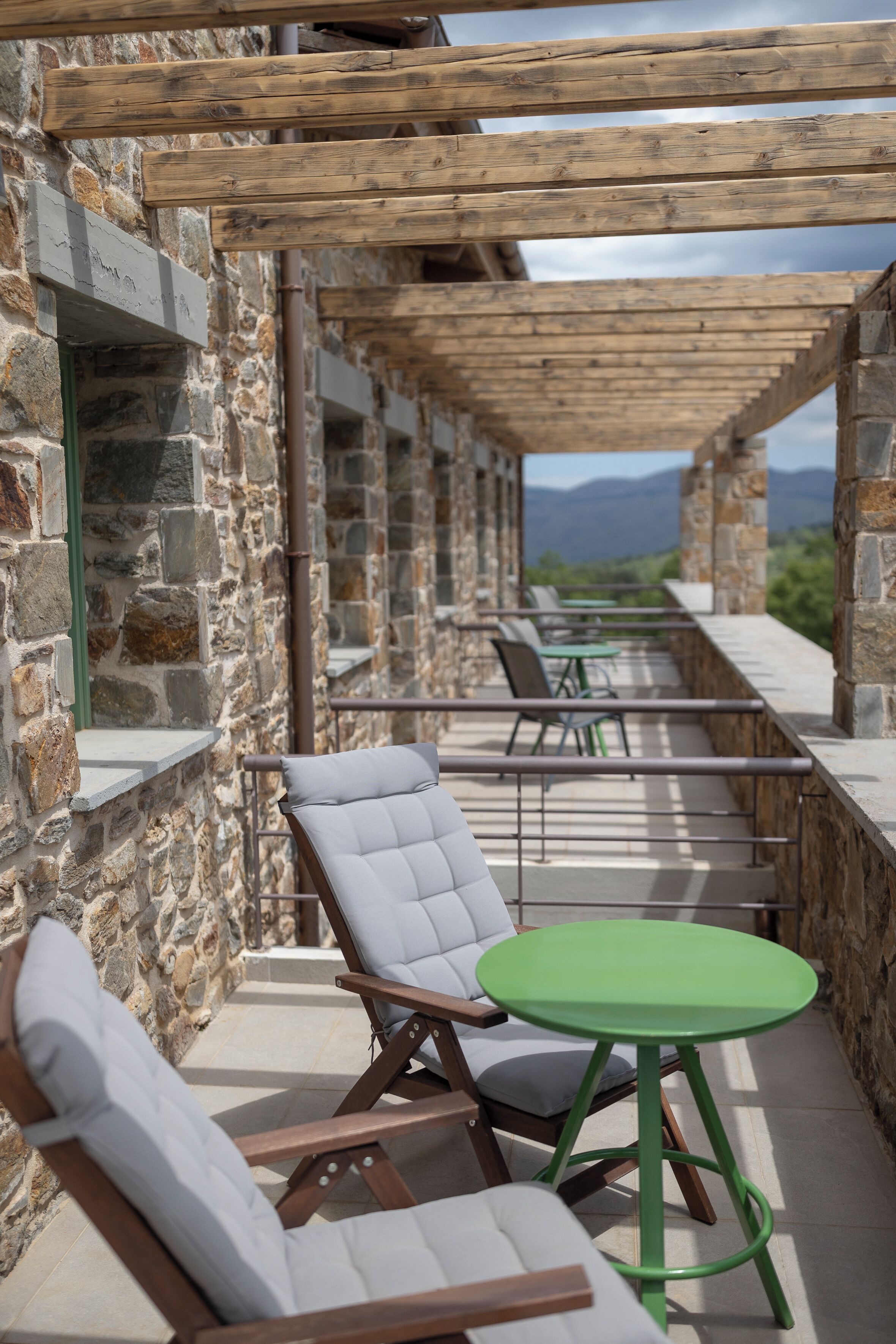 A stone building with a wooden pergola, outdoor seating with chairs and a table, and a view of mountains in the background.
