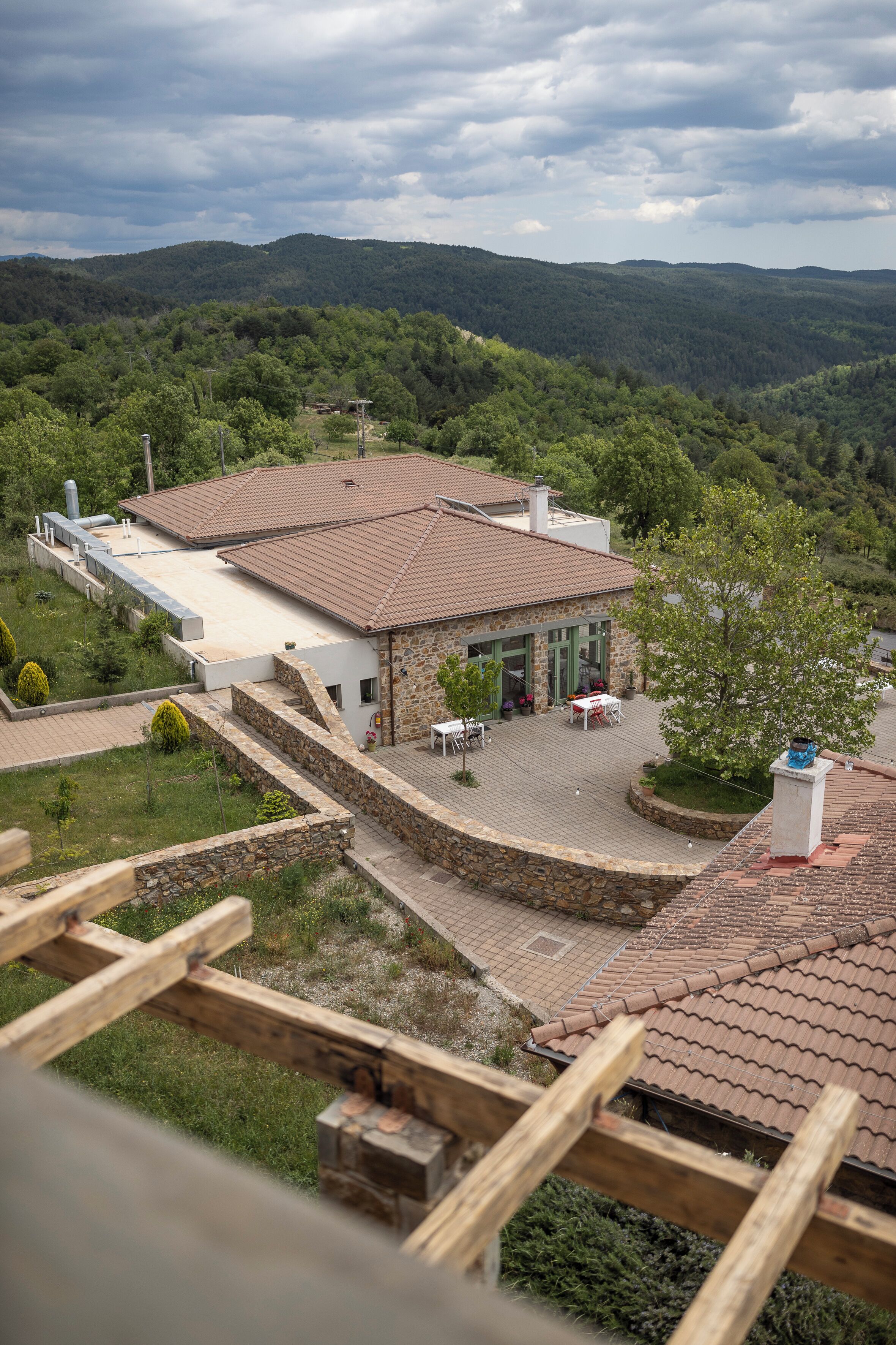 A stone building with a tiled roof, surrounded by a landscaped garden and a view of a forested valley.