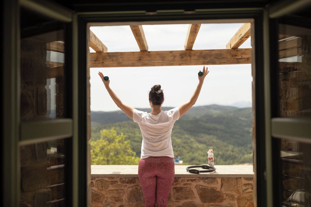 A person standing with arms raised, looking out from a wooden balcony with a stone wall, overlooking a mountainous landscape.