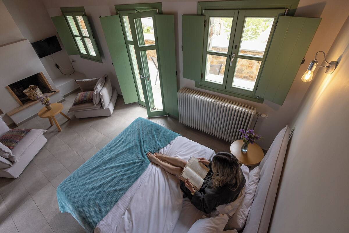 A bedroom with a bed, a reading chair, a small table, a radiator, and a window with shutters.