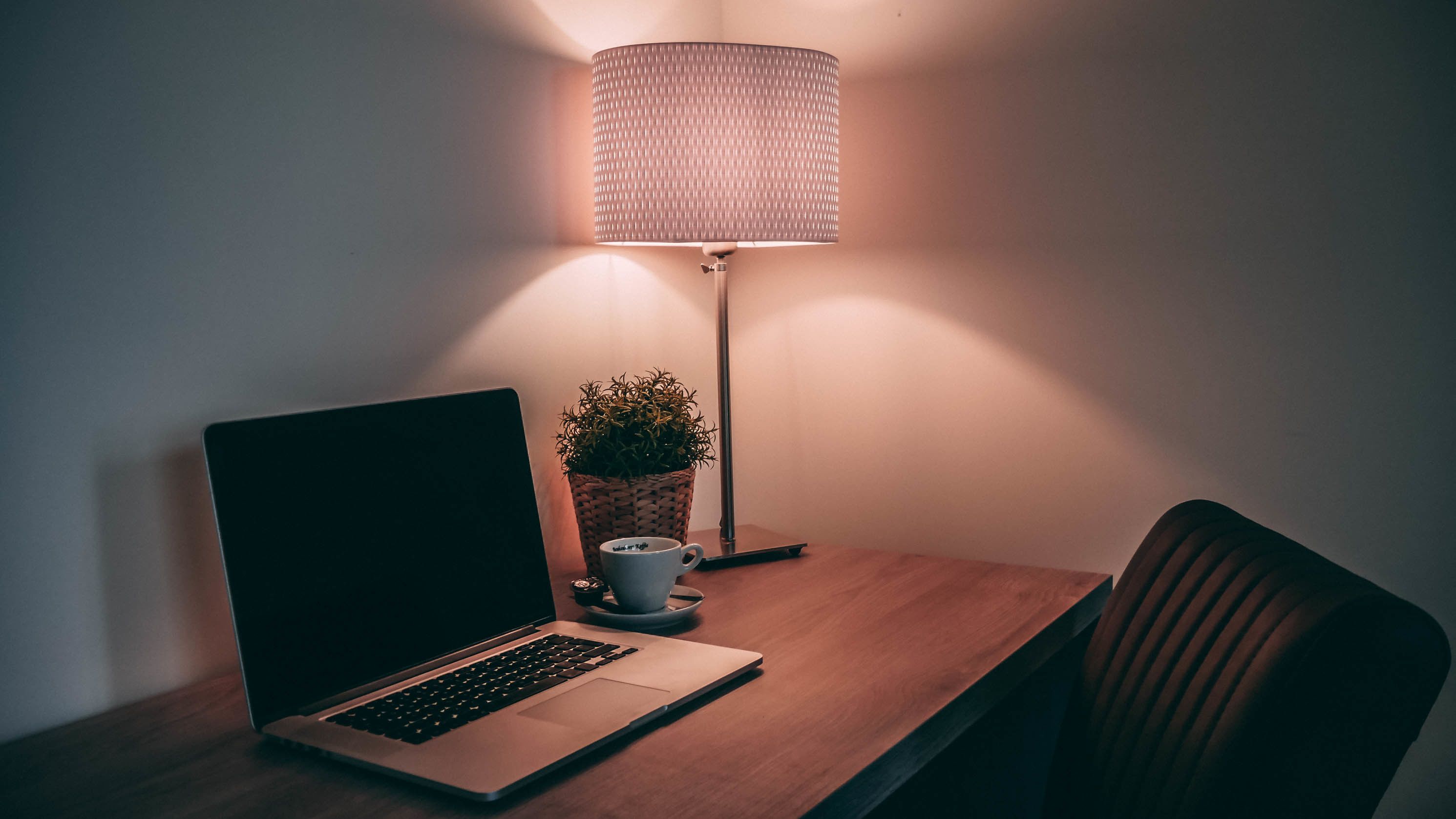 Een laptop op een houten bureau met een bloempot en een kop koffie. Een vloerlamp met een geweven kap staat op het bureau.