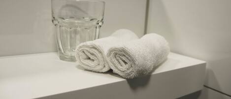Un estante blanco en el baño con un vaso transparente y dos toallas blancas enrolladas.