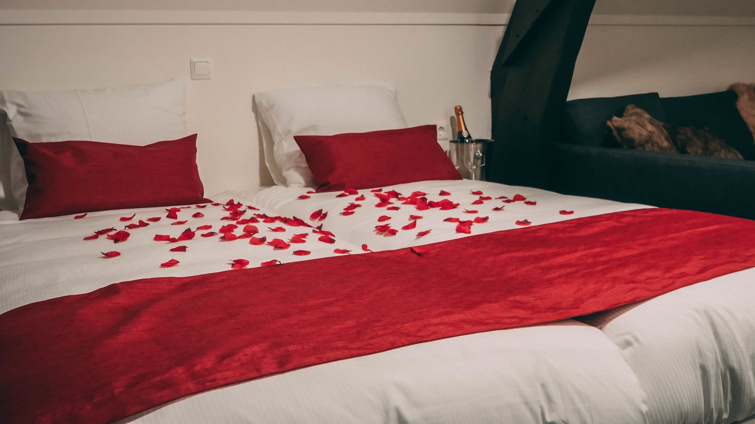 A bed with red and white bedding, a bottle of champagne, and a glass on the nightstand.