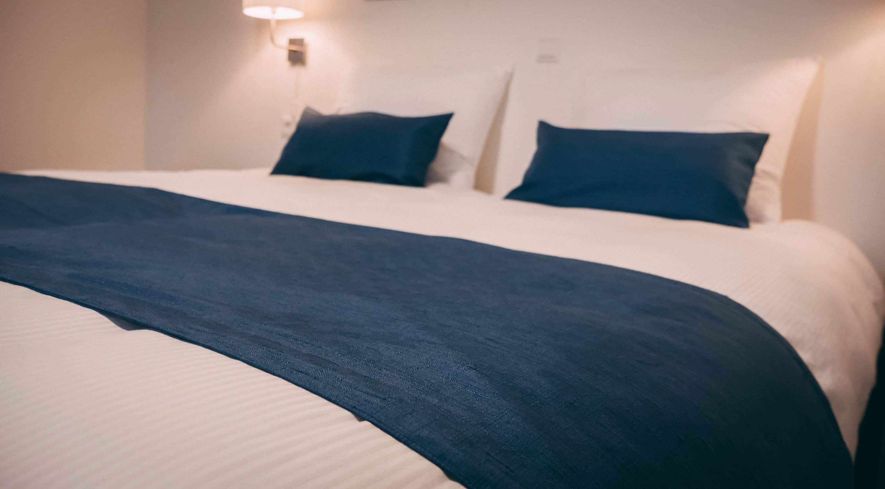 A neatly made bed with a dark blue blanket and pillows, a white headboard, and a wall-mounted light fixture.