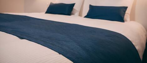 A neatly made bed with a dark blue blanket and pillows, a white headboard, and a wall-mounted light fixture.
