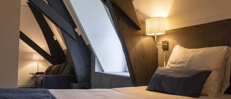 A bedroom with a sloped ceiling, a bed with white and dark pillows, a nightstand with a lamp, and a window with a view.