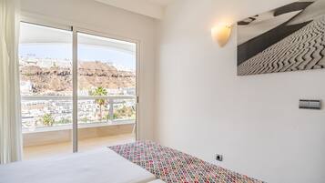 A hotel room with a large window, a bed, a patterned rug, a wall-mounted light, and a framed picture.