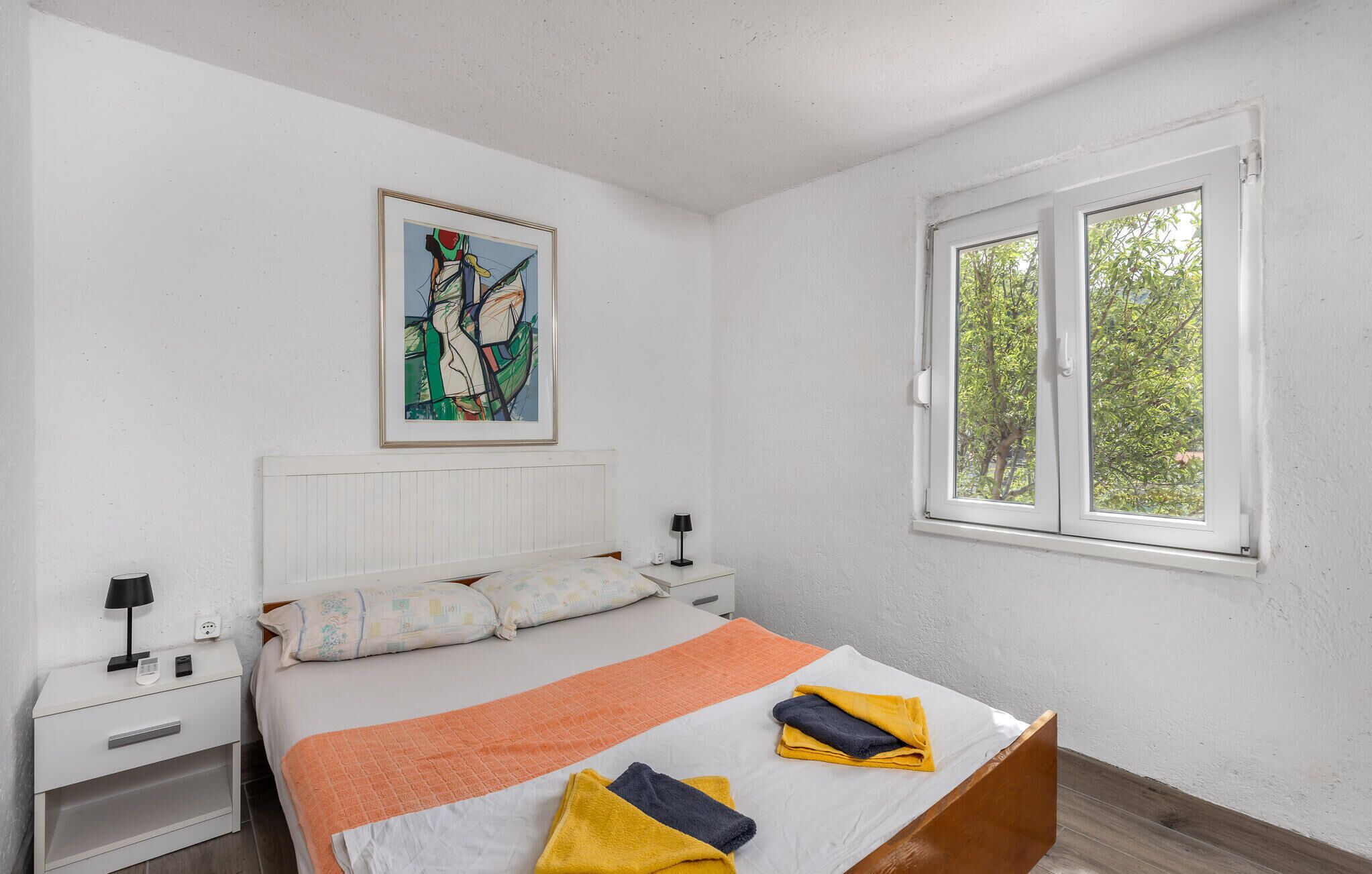 A bedroom with a bed, a nightstand, a lamp, a painting on the wall, and a window with a view of trees.