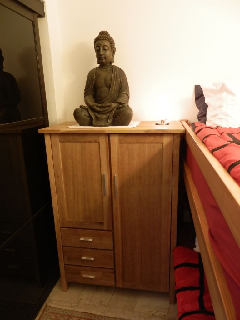 Ein Zimmer mit einem Holzkasten, einem Bett mit rot-schwarzen Streifen und einer Buddha-Statue auf dem Kasten.