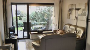 En stue med sofa, fjernsyn, bord og et vindue med udsigt til grøn vegetation.
